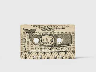 Scrimshaw in the shape of a cassette, with a drawing of 2 people warming by a campfire, in the belly of a whale with the text Do Something Crazy