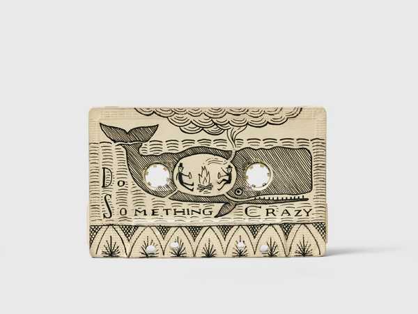 Scrimshaw in the shape of a cassette, with a drawing of 2 people warming by a campfire, in the belly of a whale with the text Do Something Crazy