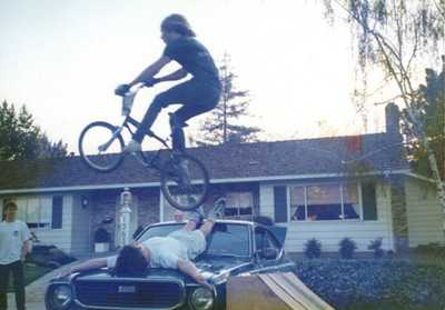 Kid jumping another kid laying on on the hood of a car with his bike