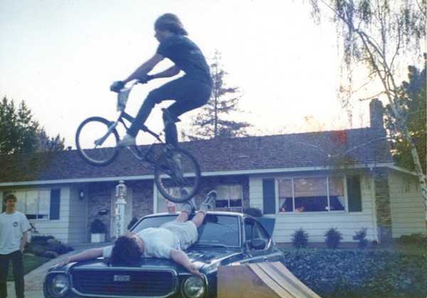 Kid jumping another kid laying on on the hood of a car with his bike
