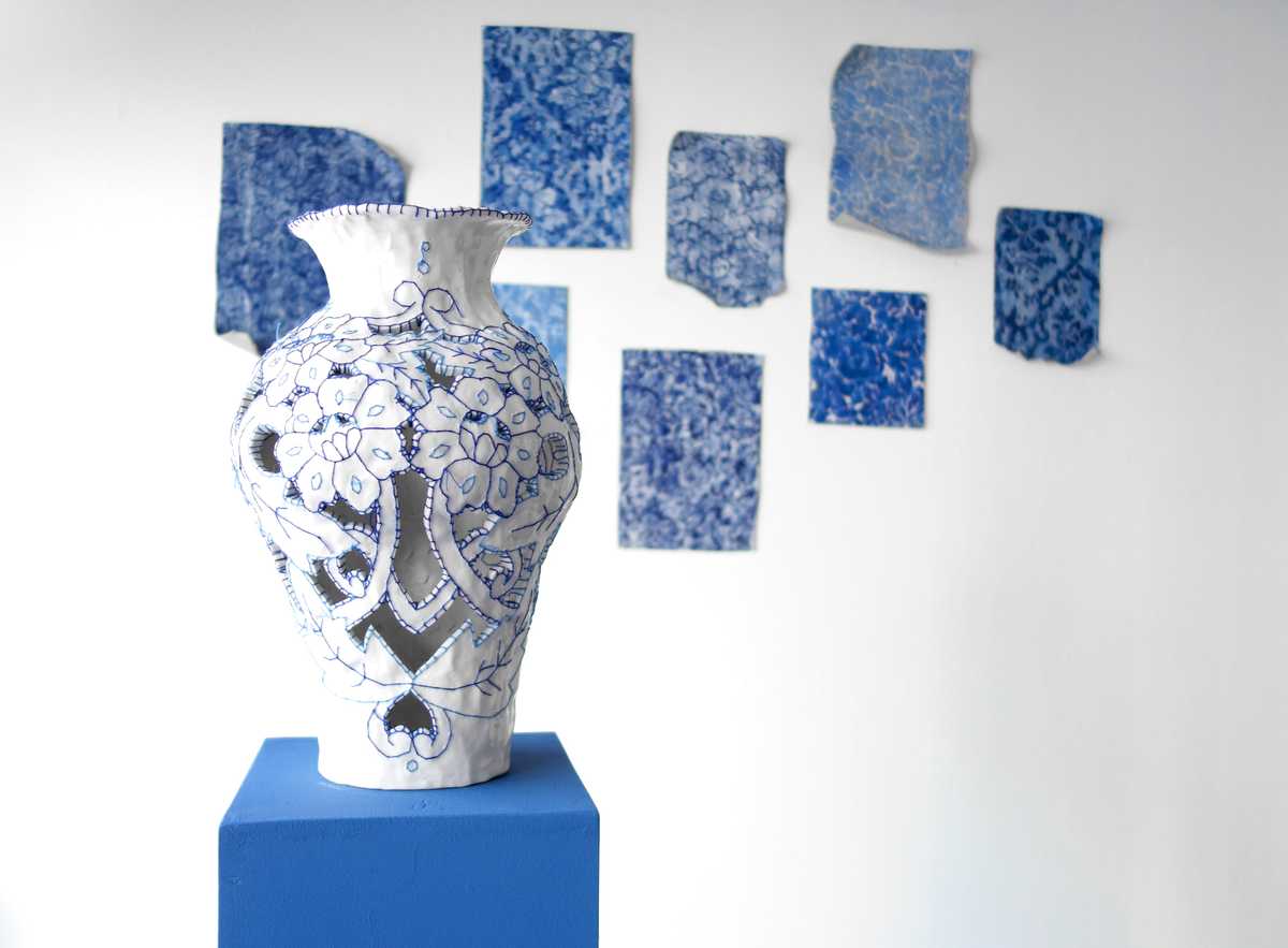 Photo of an embroidered ceramic vase with more artwork behind it on a wall