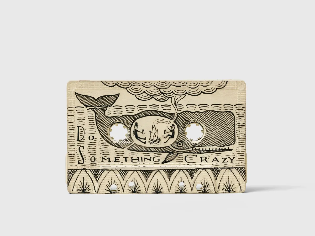 Scrimshaw in the shape of a cassette, with a drawing of 2 people warming by a campfire, in the belly of a whale with the text Do Something Crazy